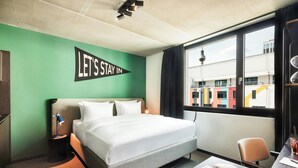 A hotel room with a bed, a desk, a chair, a small table, a lamp, a magazine, and a window with a view of buildings. - The Social Hub Berlin (Berlin)