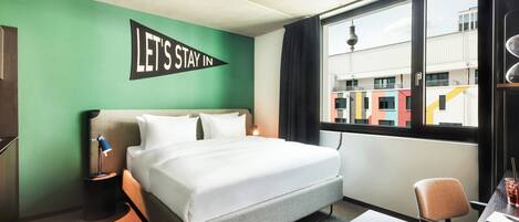 Habitación de hotel con cama, escritorio, silla, mesita, lámpara, revista y ventana con vista a edificios.