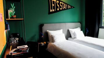 Une chambre d’hôtel avec un lit, un bureau doté d’une lampe, une étagère avec une plante et un panneau indiquant : « Restons à l’hôtel ».