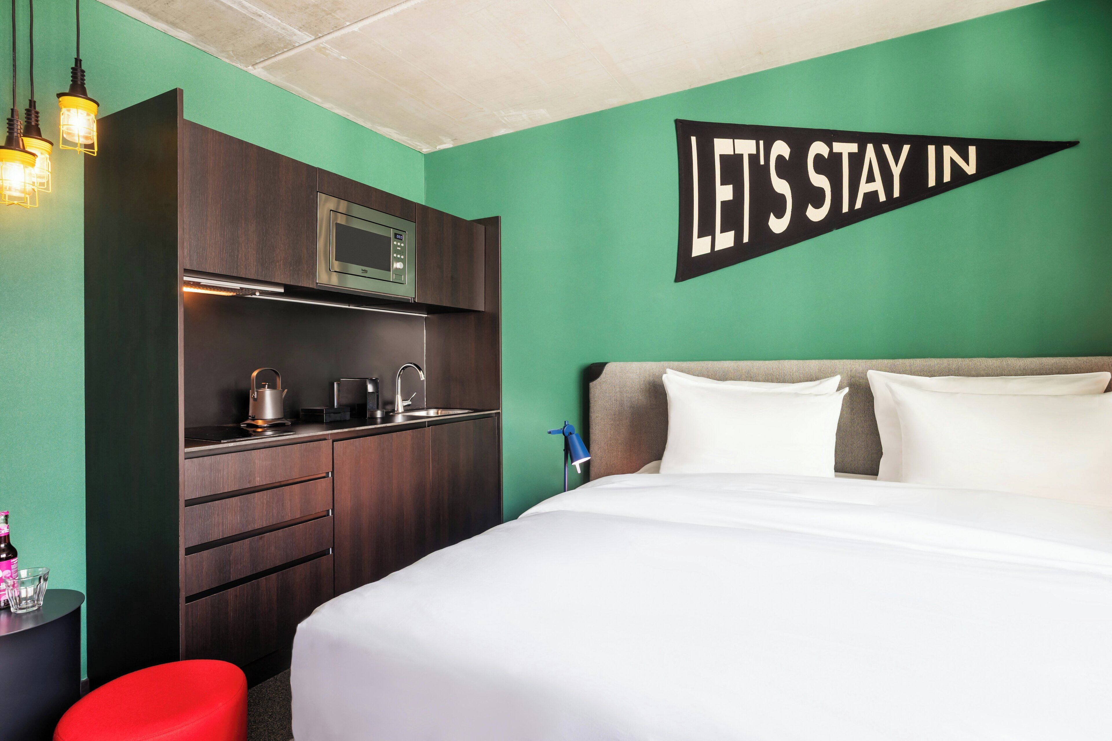 A hotel room with a bed, a kitchenette, and a wall sign that says 'LET'S STAY IN'.