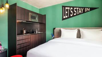 A hotel room with a bed, a kitchenette, and a wall sign that says 'LET'S STAY IN'.