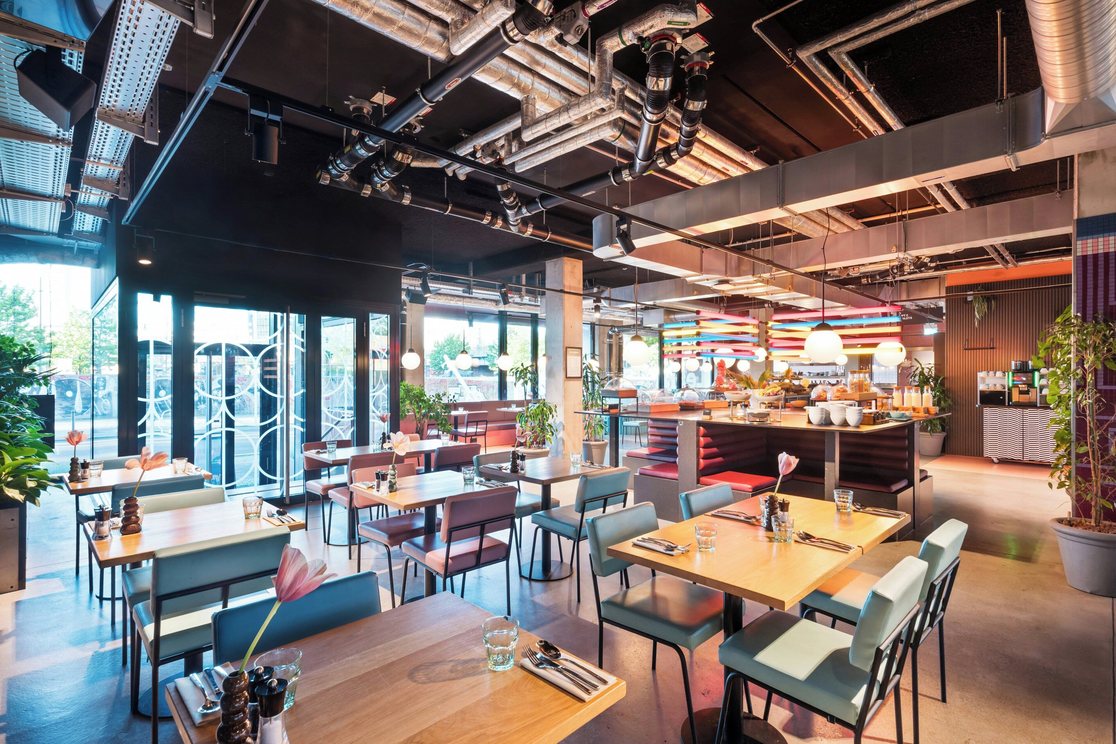 A modern restaurant interior with exposed ductwork, wooden tables, and chairs, and a visible kitchen area.