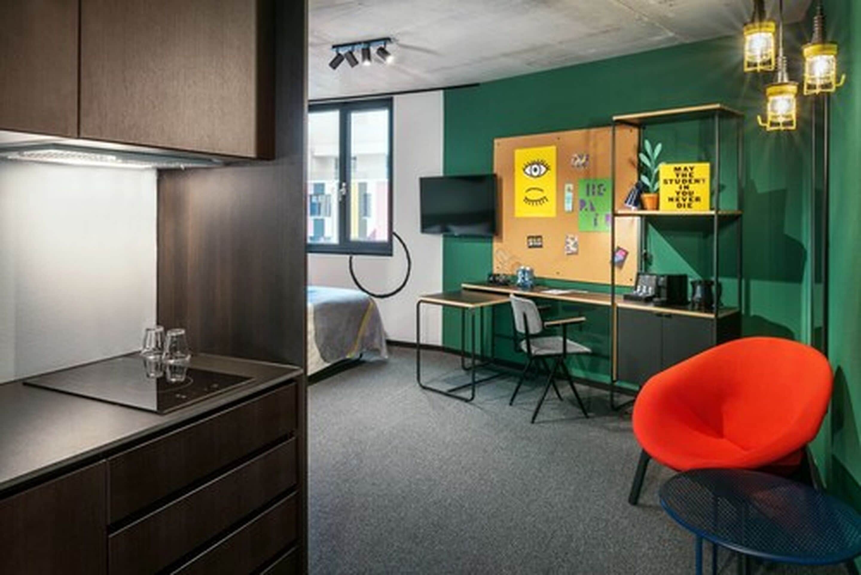 Un espacio de oficina moderno con kitchenette, un escritorio y una silla, una silla naranja brillante y un televisor en la pared.