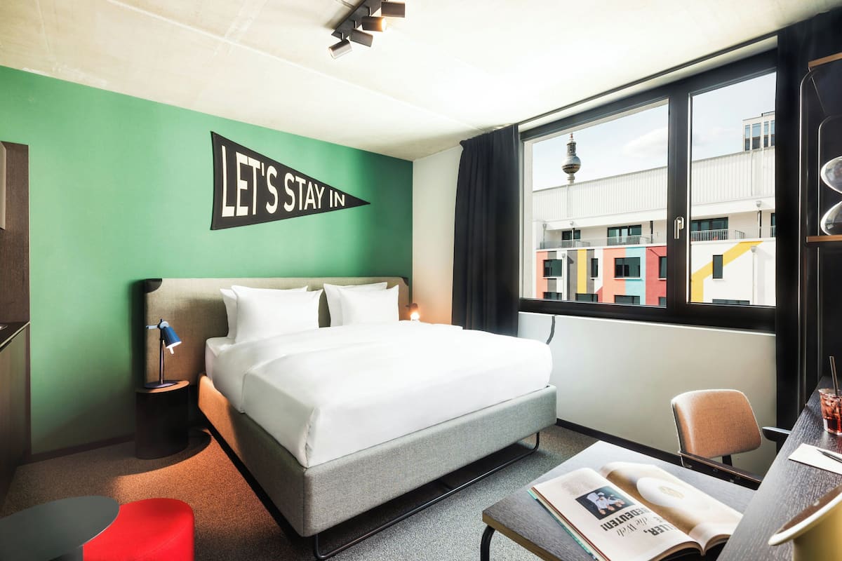 A hotel room with a bed, a desk, a chair, a small table, a lamp, and a window with a view of buildings.