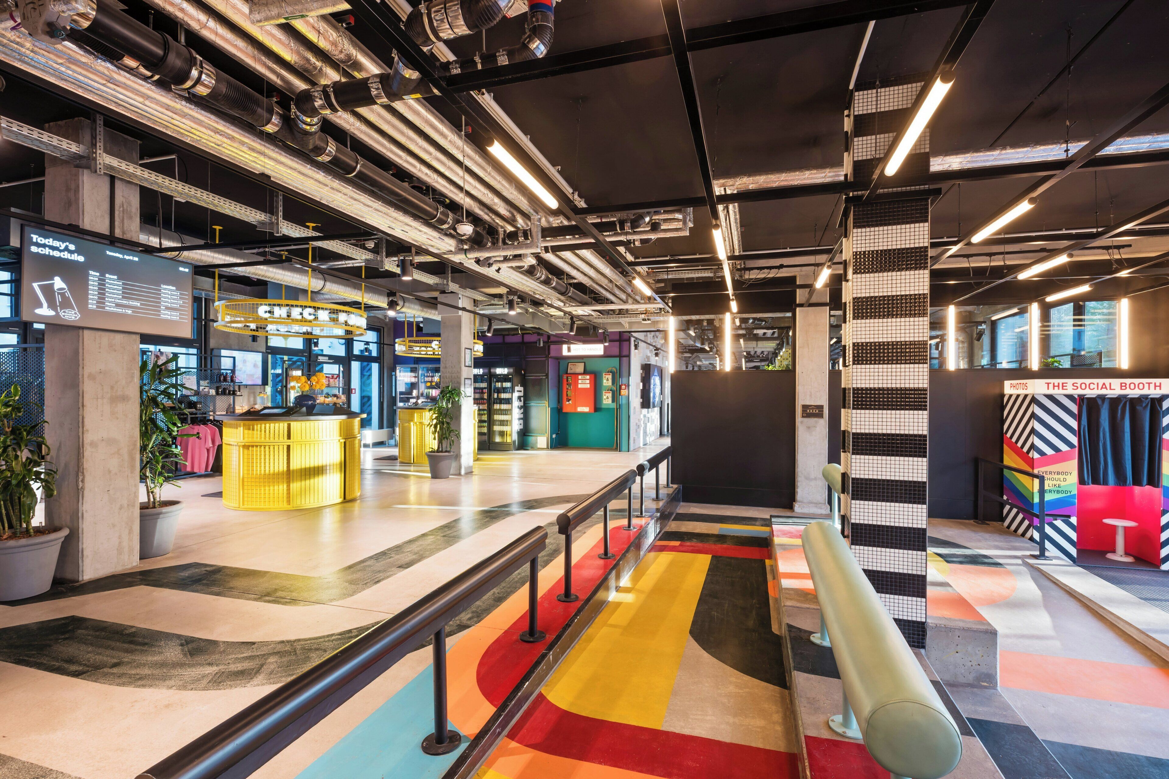 A modern indoor space with exposed ductwork, a digital display board, a yellow counter, and a colorful floor design.