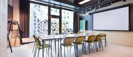 A conference room with a long table, chairs, a projector screen, and a large window with a view of a building.