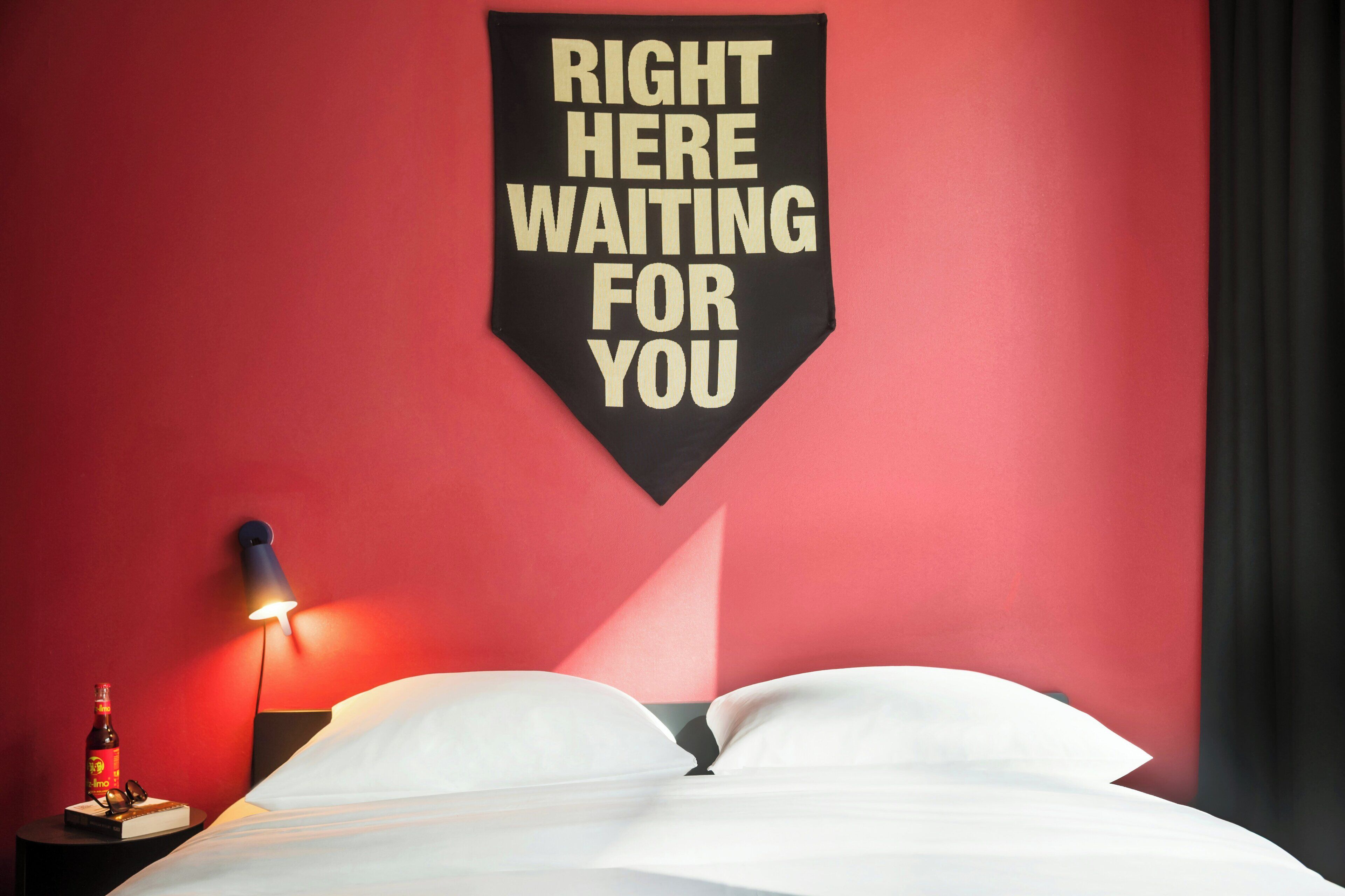 A hotel room with a bed, a wall-mounted sign, a bedside table with a bottle and glasses, and a wall lamp.