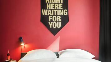 A hotel room with a bed, a wall-mounted sign, a bedside table with a bottle and glasses, and a wall lamp.