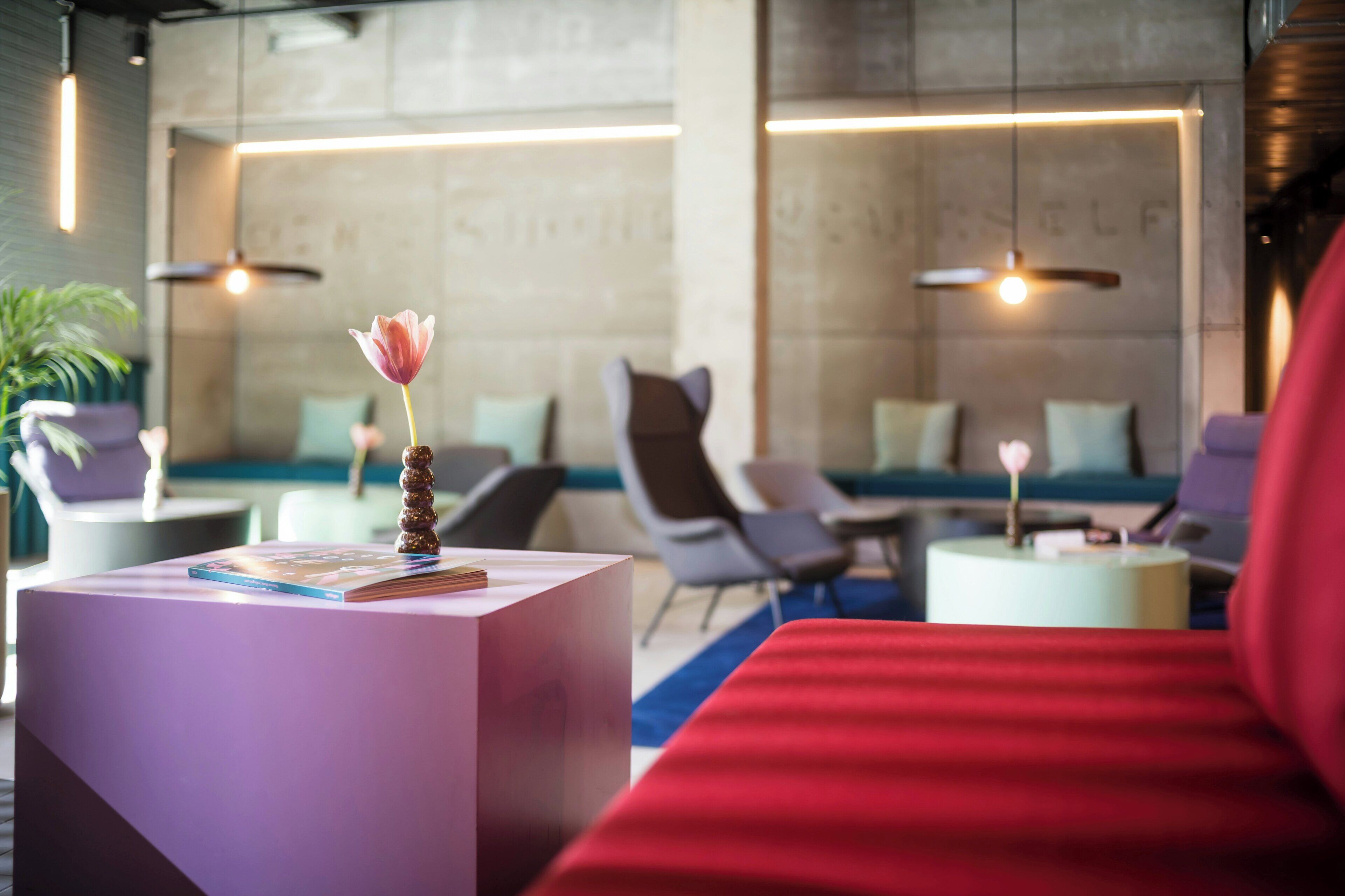 Eine moderne Hotelhalle mit einem roten Sofa, einem weißen Couchtisch mit Buch und Vase sowie einer Sitzecke mit Stühlen und einem kleinen Tisch.