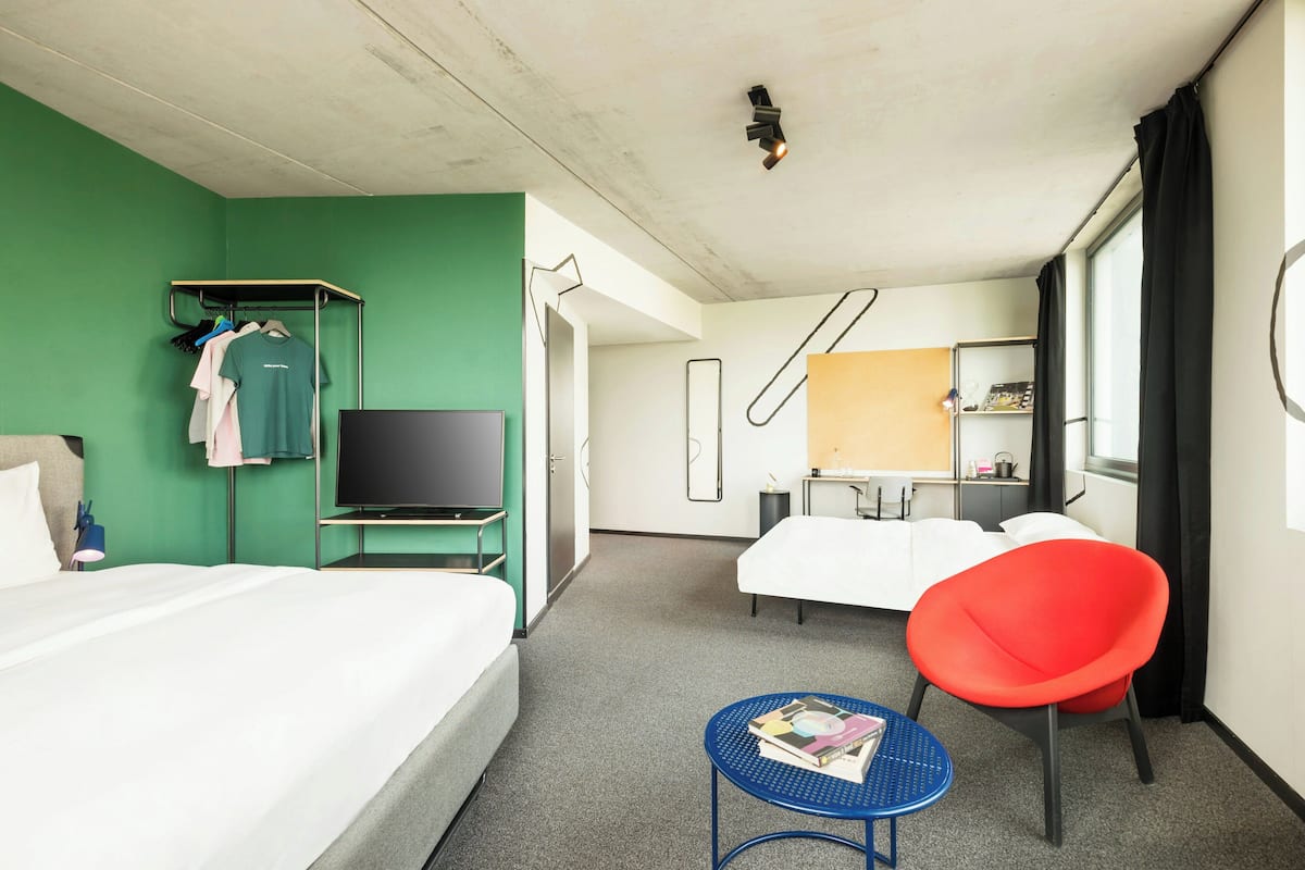 A hotel room with a bed, a TV, a red chair, a small table, and a green wall.