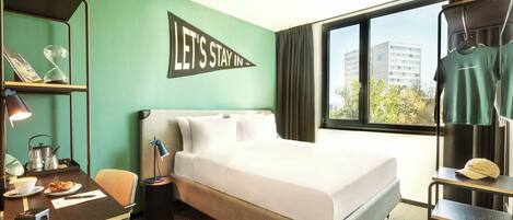 A hotel room with a bed, a desk with a chair, a small table with a lamp, a shelf with a clock, and a window with a view of buildings.