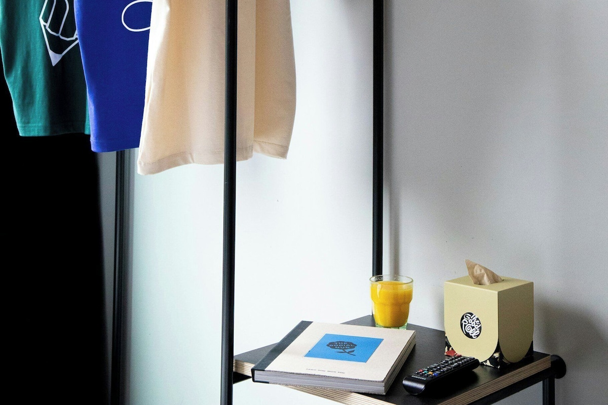 A clothing rack with three t-shirts, a remote control, a book, a tissue box, and a glass of orange juice on a table.
