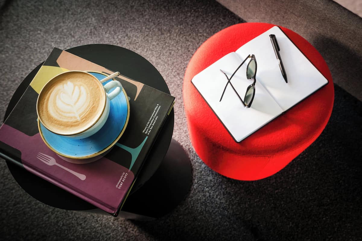 A table with a coffee cup, a book, and a red ottoman with sunglasses and a pen on it.