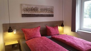 Un lit double avec une literie rouge, deux tables de chevet, une œuvre d’art accrochée au mur et une fenêtre avec des rideaux.