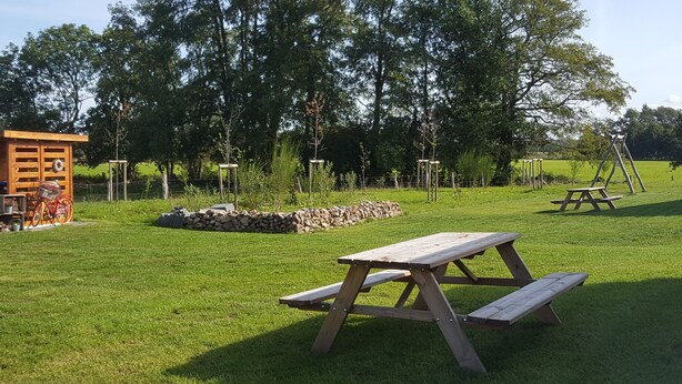 Un espace pique-nique équipé de bancs en bois, d’un muret en pierre et d’une structure en bois où est adossé un vélo.