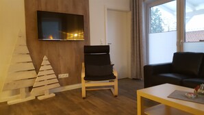 Una sala de estar con piso de madera, un sofá negro, una silla de madera, una mesita, un televisor montado en la pared y un árbol de Navidad.
