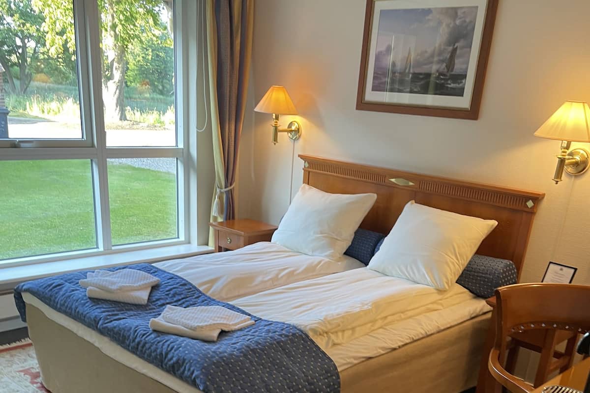 Un lit bien fait, avec une couverture bleue et des oreillers blancs, une table de chevet en bois avec une lampe, un tableau encadré au mur et, par la fenêtre, une vue sur un espace verdoyant.