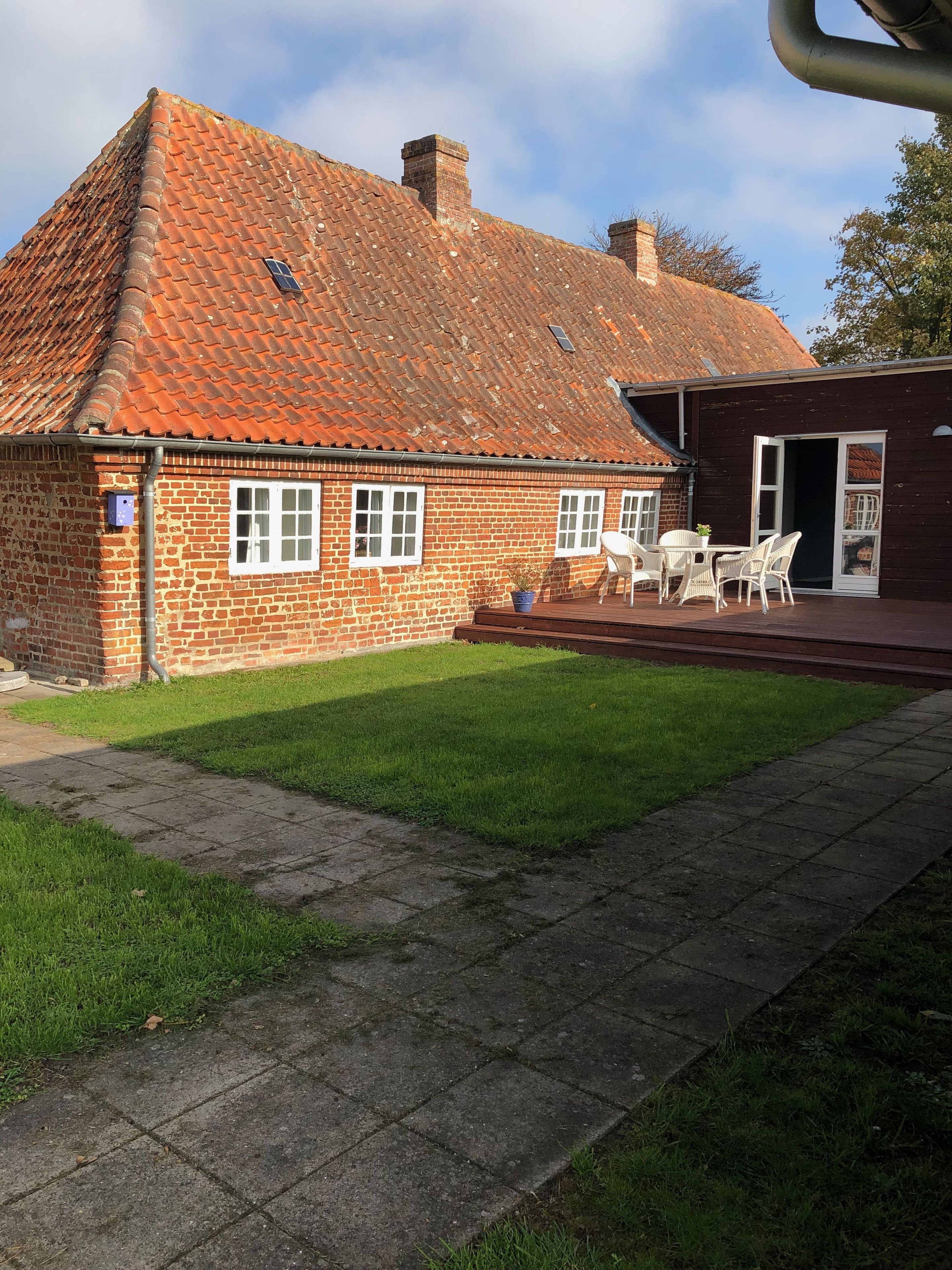 Une maison en briques avec un toit en tuiles, une cour gazonnée et une terrasse aménagée avec du mobilier d’extérieur.
