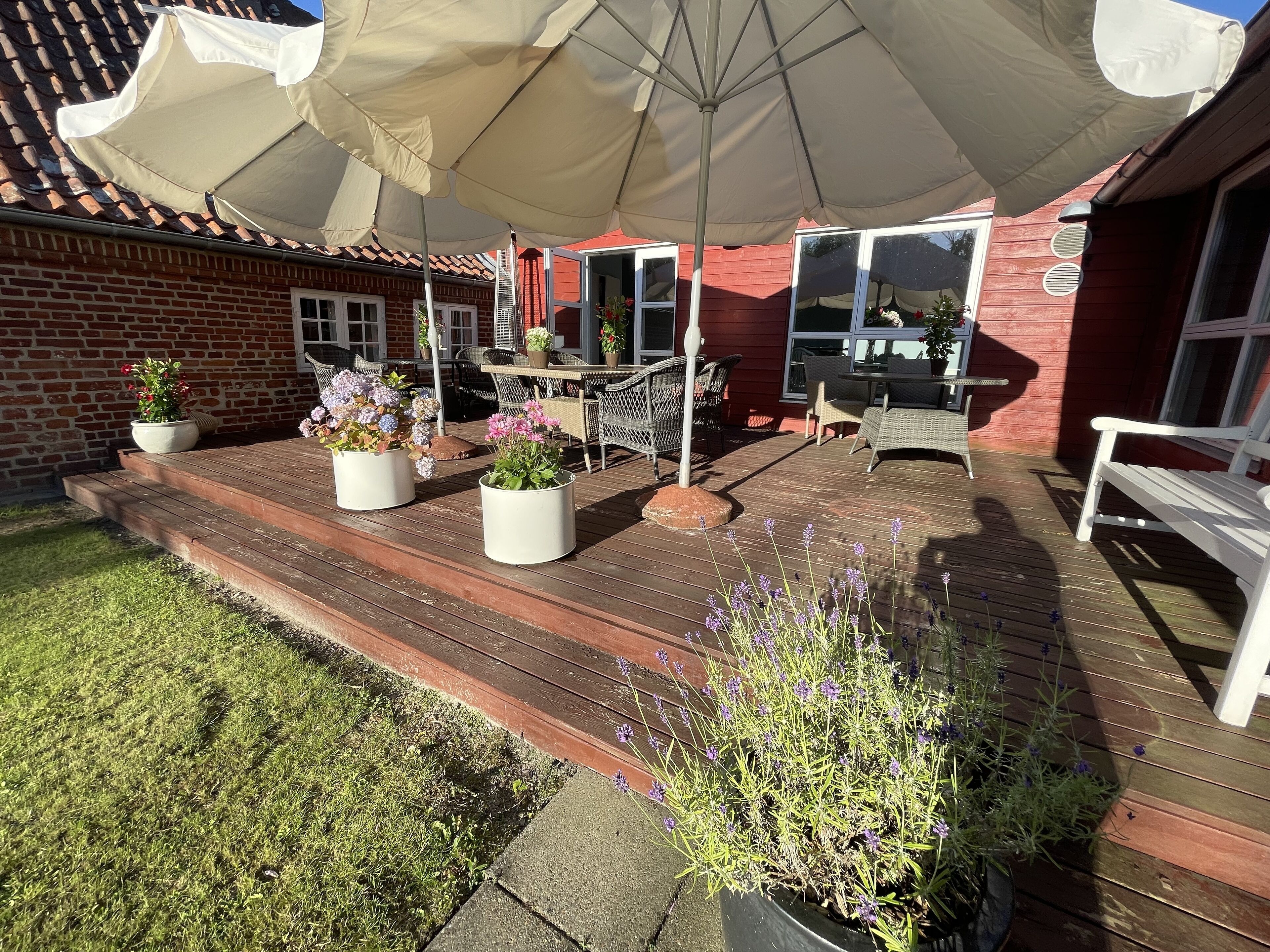 Una terraza de madera con mobiliario exterior, plantas en macetas y un amplio paraguas que da sombra.