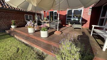 Una terraza de madera con mobiliario exterior, plantas en macetas y un amplio paraguas que da sombra.