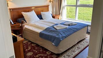 A hotel room with a large bed, a wooden headboard, two bedside lamps, a framed picture on the wall, and a view of a green lawn through a window.
