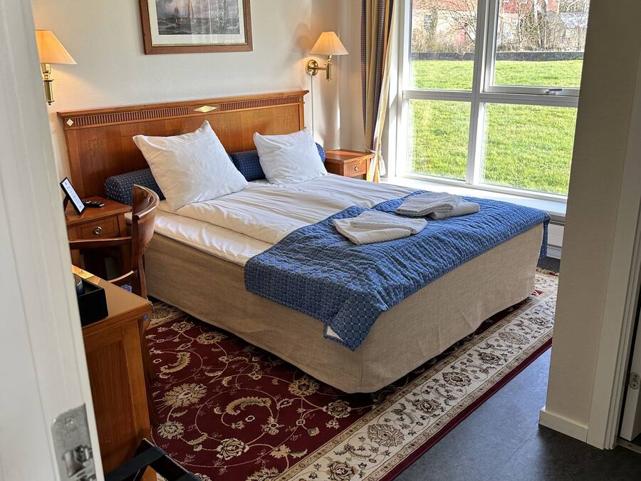 A hotel room with a large bed, a wooden headboard, two bedside lamps, a framed picture on the wall, and a view of a green lawn through a window.