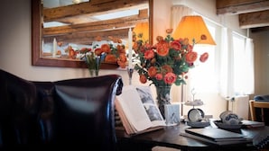 Una habitación acogedora con techo de madera, un espejo que refleja la habitación, una mesa con un libro, un florero con flores y una lámpara.