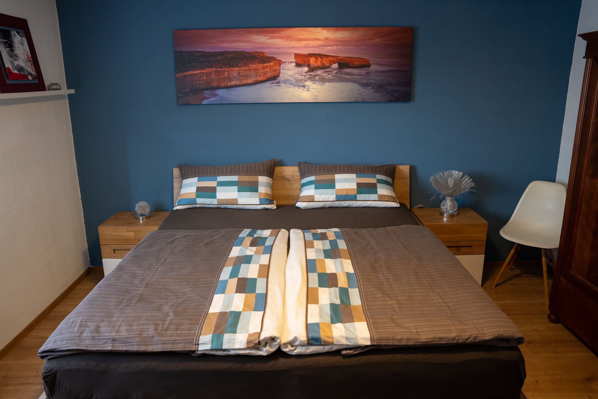 Un lit double avec une couvre-lit à carreaux, deux tables de chevet avec des lampes, une chaise blanche et un tableau représentant un coucher de soleil accroché au mur.