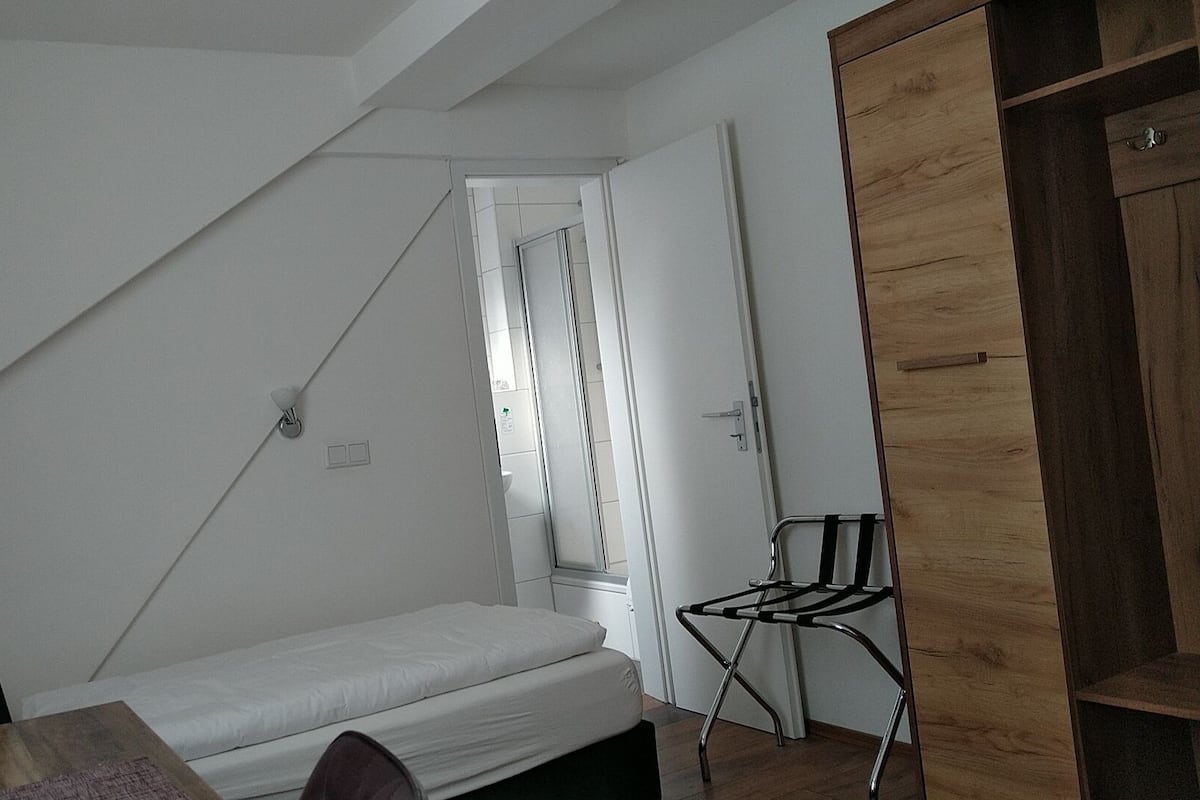 A small, sparsely furnished room with a bed, a chair, and a wooden wardrobe.