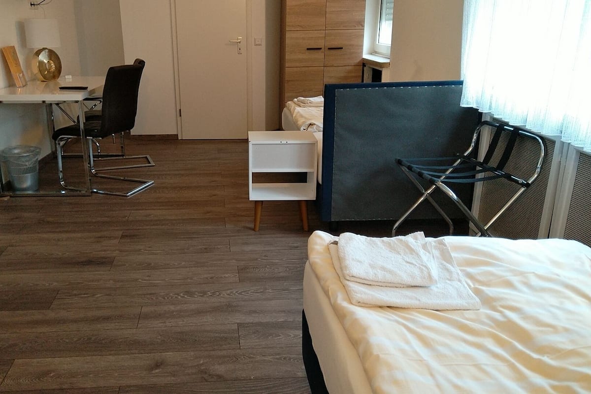 A bedroom with a bed, a desk, and a chair. There is a window with curtains and a radiator.