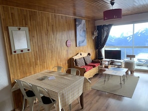 Un salon chaleureux avec un mur en bois, un canapé, une télévision, une table à manger et une vue sur les montagnes par la fenêtre.