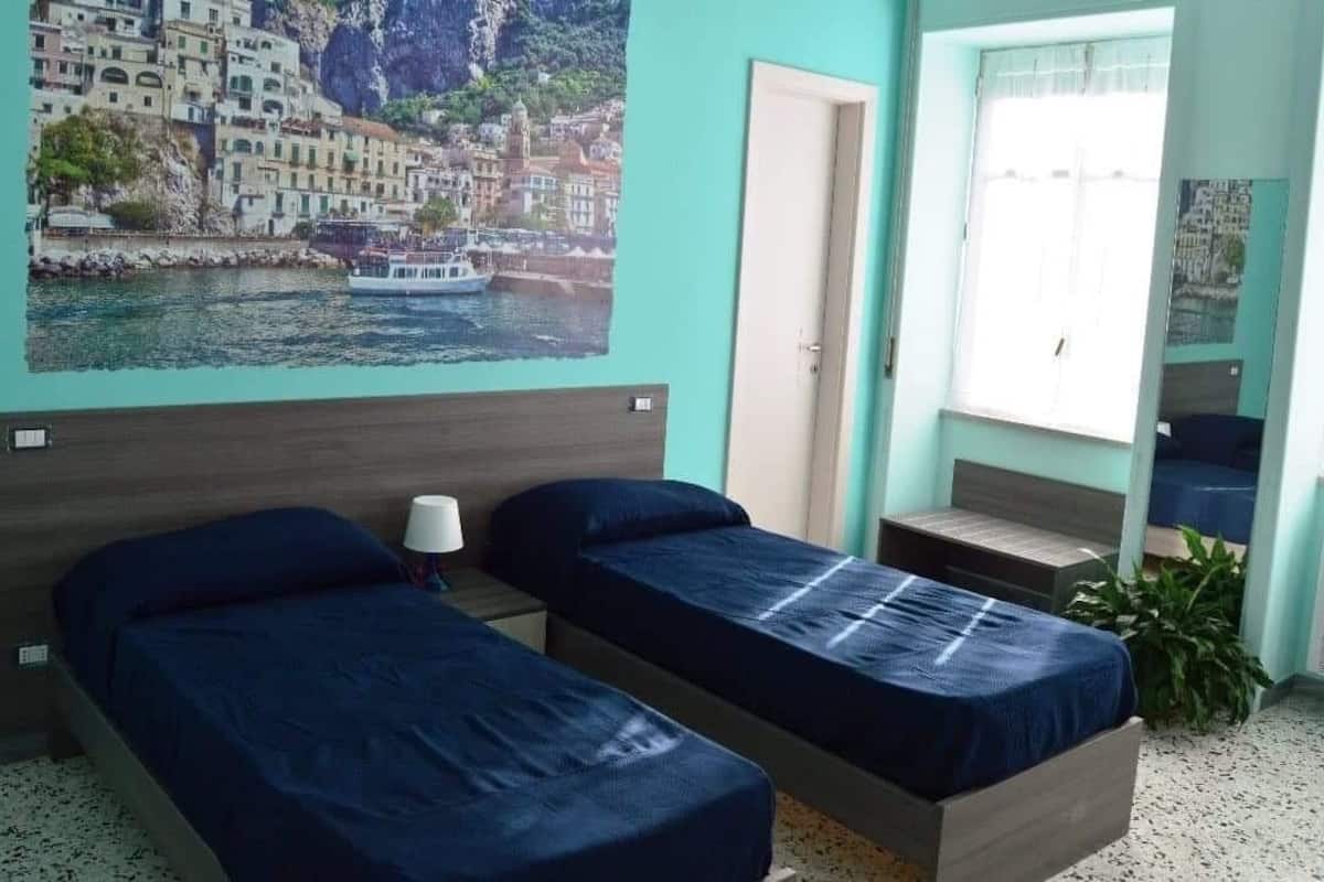 A room with two beds, a window with curtains, a wall-mounted air conditioner, and a poster of a coastal town.