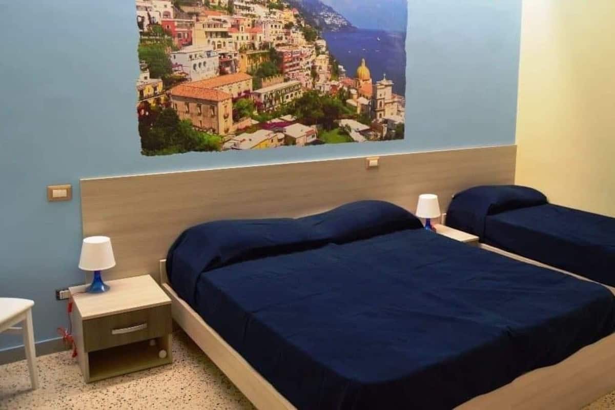 A room with two beds, a nightstand with a lamp, and a wall-mounted picture of a coastal town.