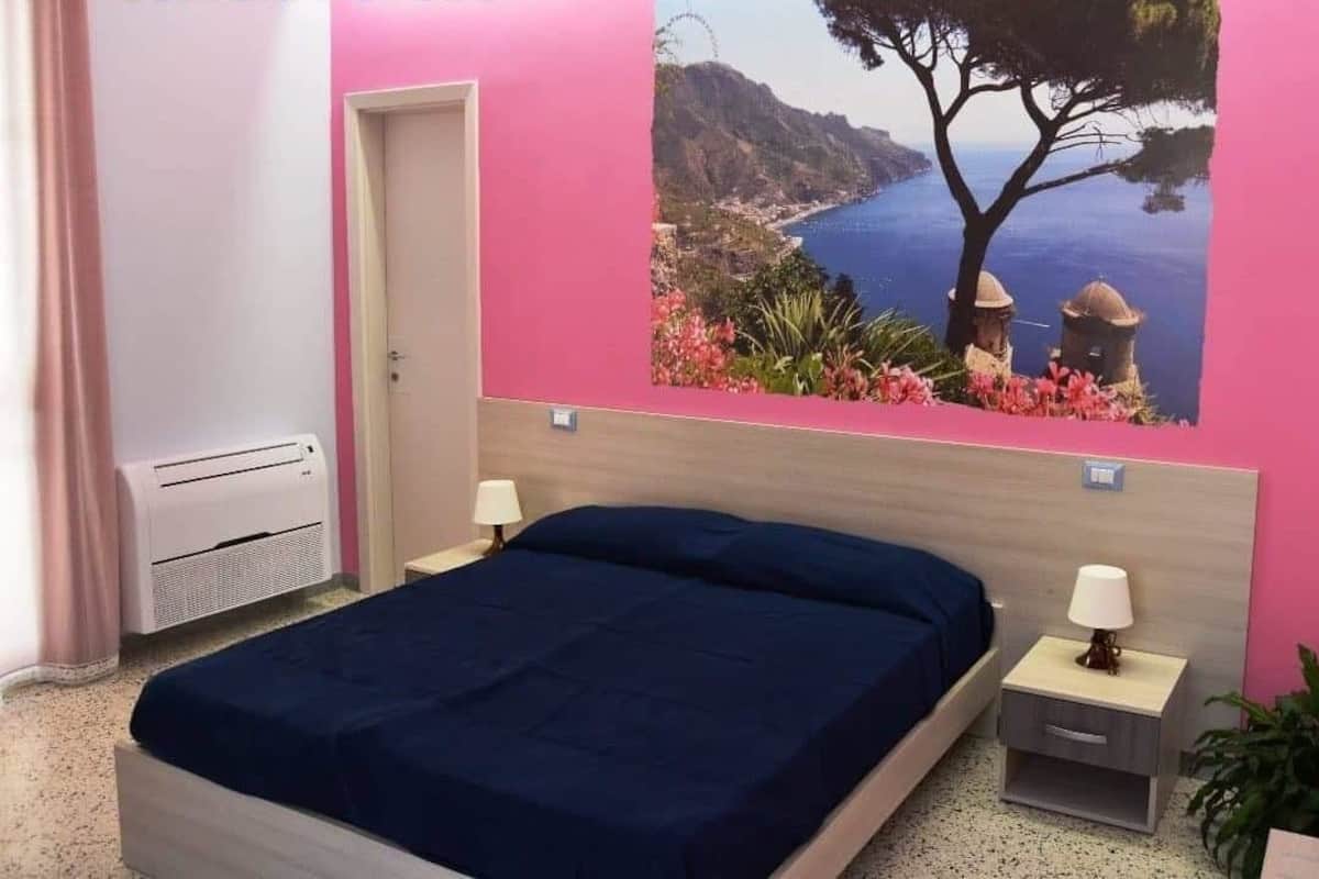 A bedroom with a bed, bedside tables, a wall-mounted air conditioner, and a large scenic painting.