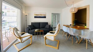 Una sala de estar moderna con un sofá, dos sillones, una mesa de comedor con sillas, una zona de cocina y un balcón con vistas a edificios.