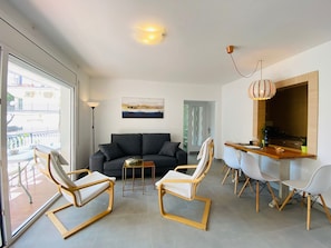 A modern living room with a sofa, two armchairs, a dining table with chairs, a kitchen area, and a balcony with a view of buildings.