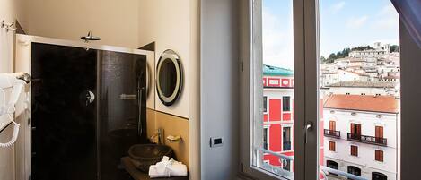 Een badkamer met een douche met glazen wand, een wastafel en een spiegel. Een raam biedt uitzicht op een stadsgezicht met gebouwen.