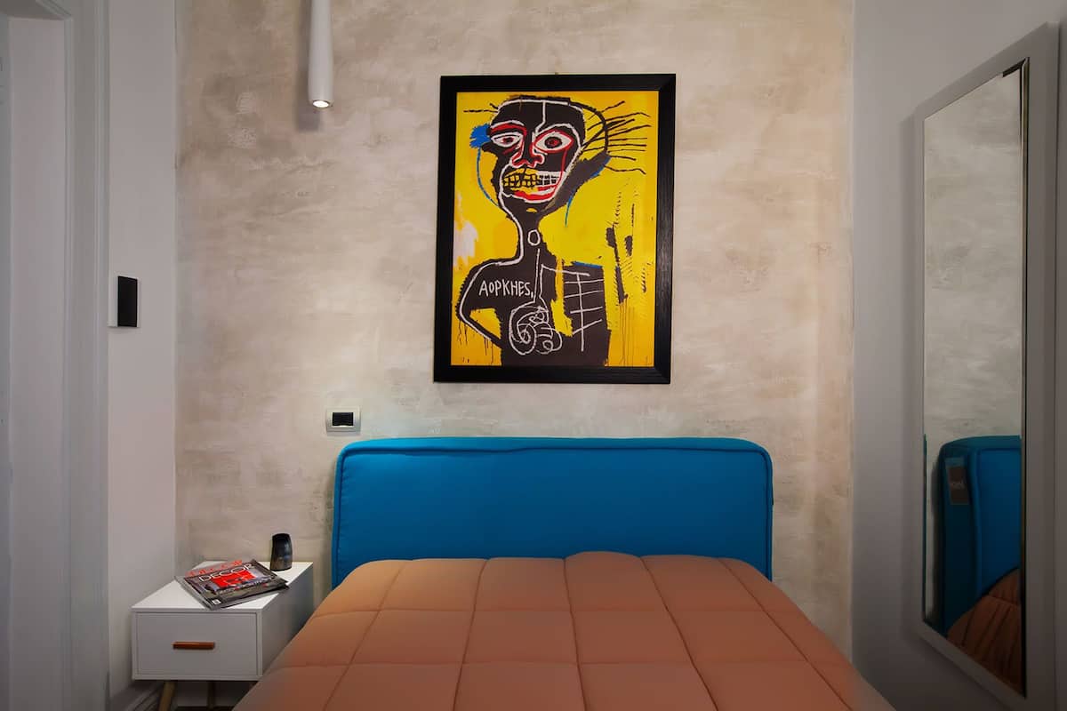 A bedroom with a blue headboard, a bed with an orange mattress, a small bedside table with a magazine, and a framed abstract painting on the wall.