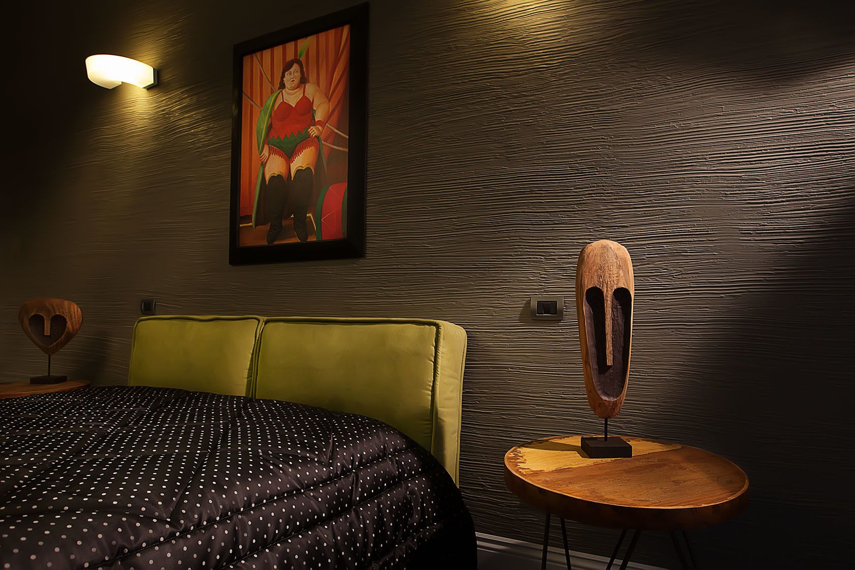 A hotel room with a bed, a yellow headboard, a round wooden table, and a wall-mounted light fixture.