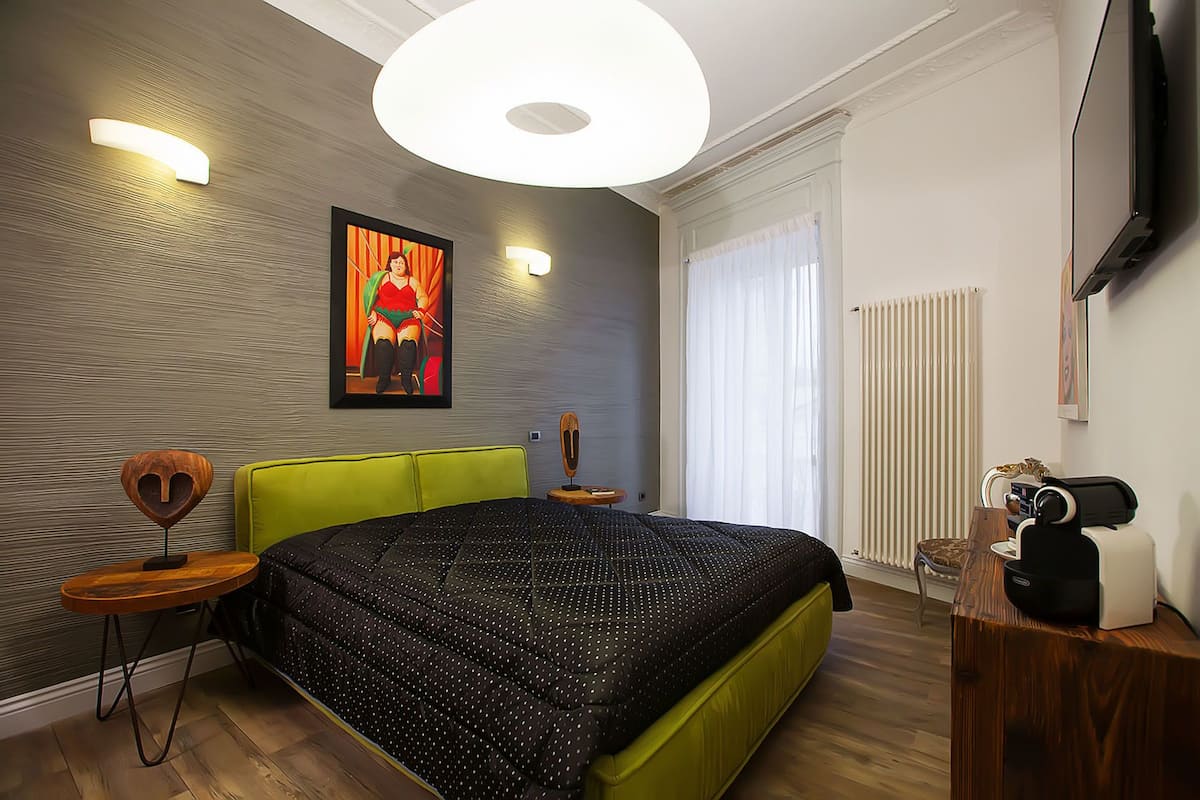 A modern bedroom with a green upholstered headboard, a large bed with a polka dot duvet, a wooden side table, a wall-mounted TV, and a radiator.