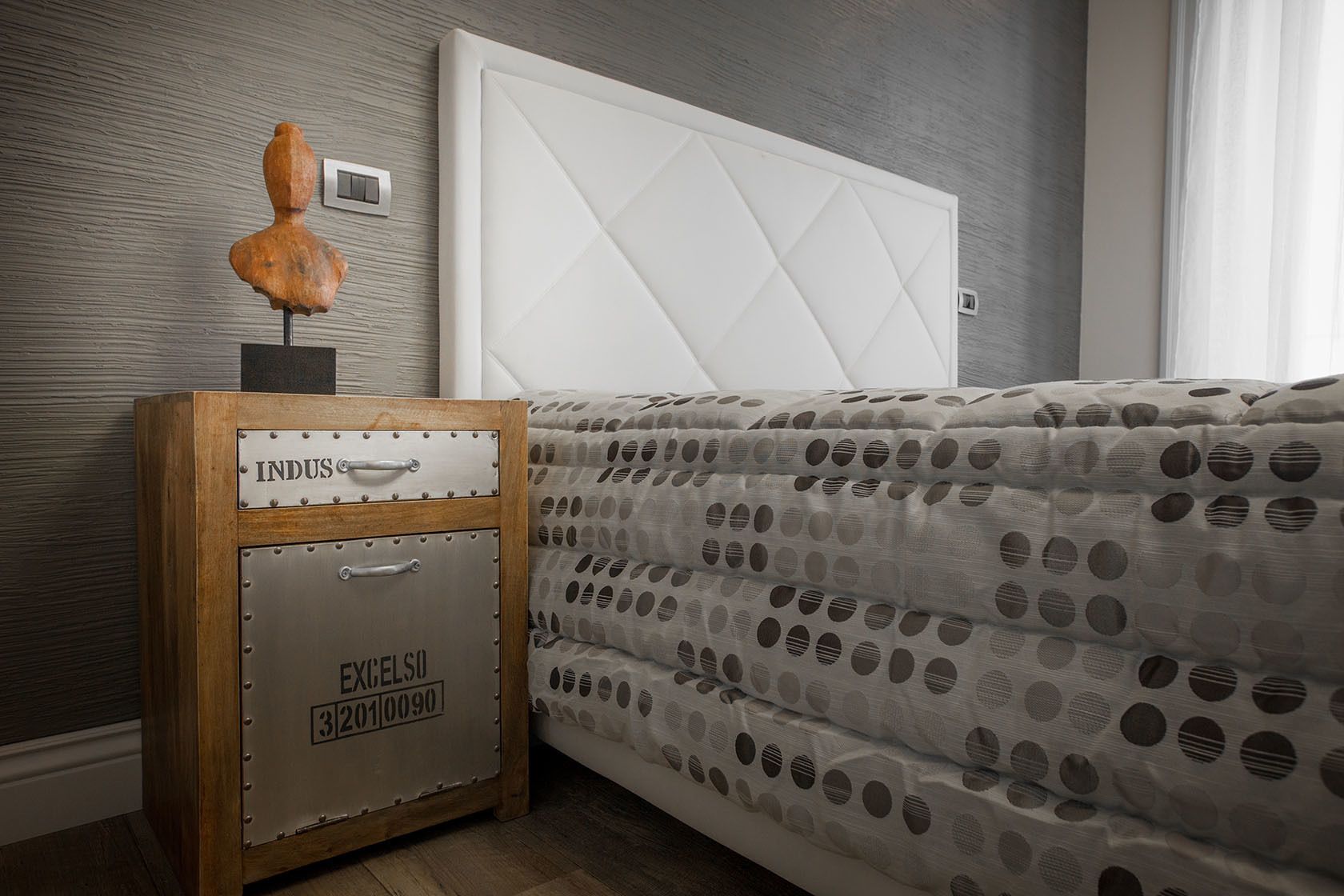 A bedroom with a bed, a nightstand with a vintage suitcase, and a wall-mounted light switch.