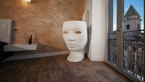 Una habitación con suelo de madera, pared blanca con textura y una consola blanca con un florero, además de una gran escultura de busto blanco.