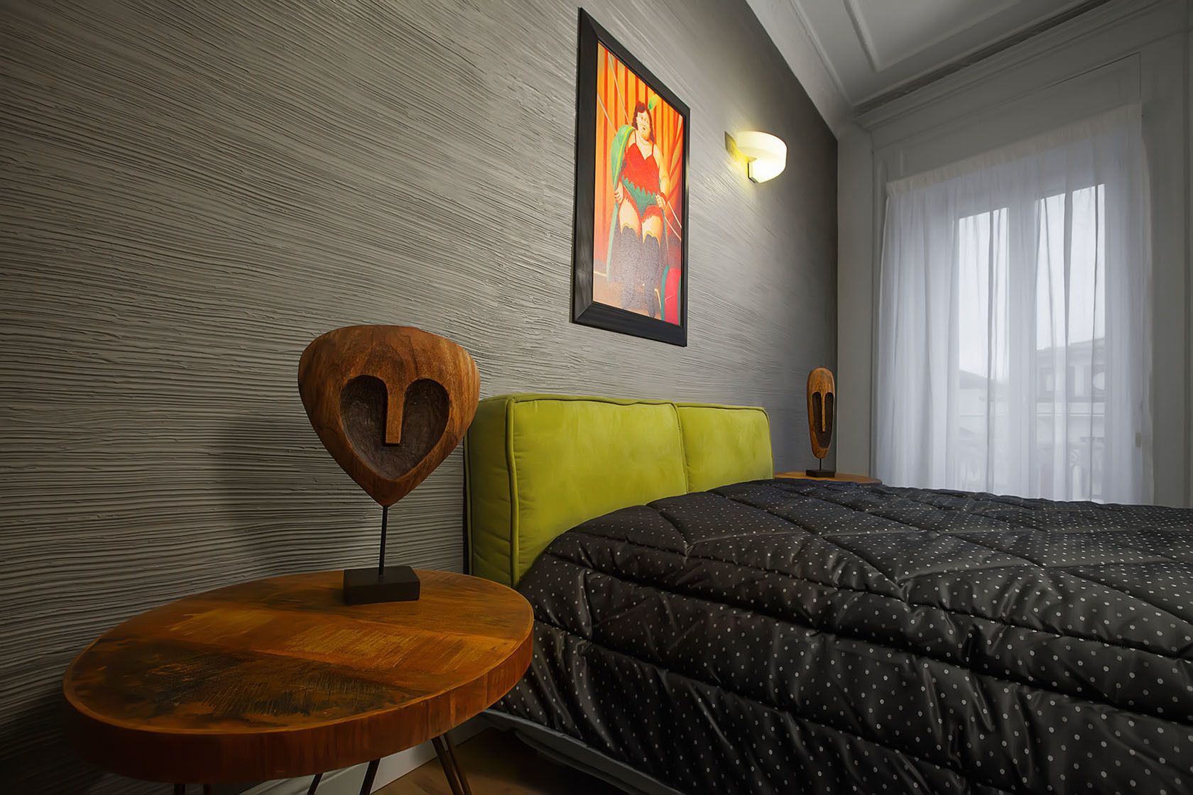Un dormitorio con cama, una mesa redonda de madera, cabecera tapizada de verde, una lámpara de pared y un cuadro enmarcado.