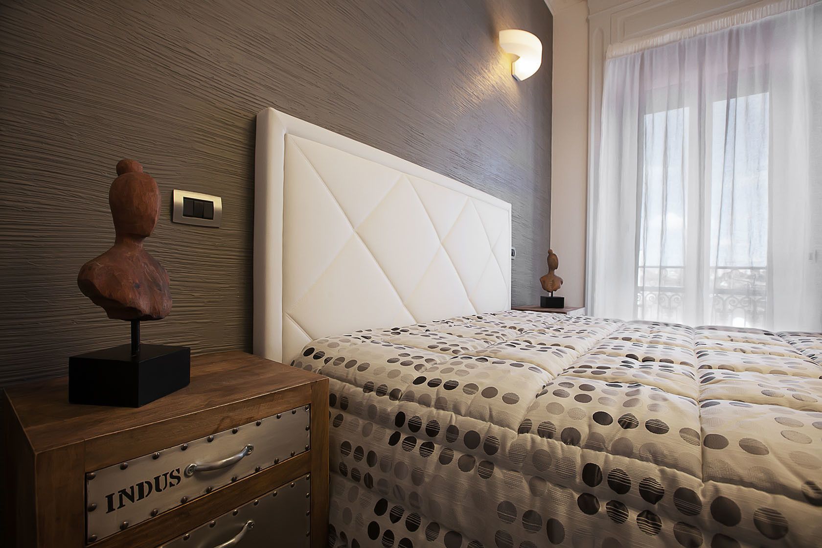 Un dormitorio con una cama grande, una mesita de noche de madera con cajones, una lámpara de pared y una ventana con cortinas traslúcidas.