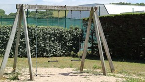 Un ensemble de balançoires en bois, installé dans un espace herbeux, avec une clôture verte et une haie en arrière-plan.