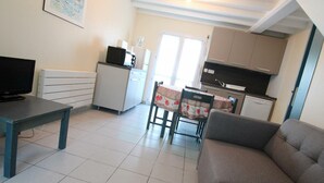 Un salon comprenant un canapé, un téléviseur, une petite table et un coin cuisine avec une table à manger et des chaises.