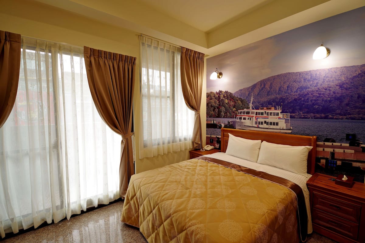 A hotel room with a large bed, a nightstand, a lamp, and a scenic mural of a boat on water.