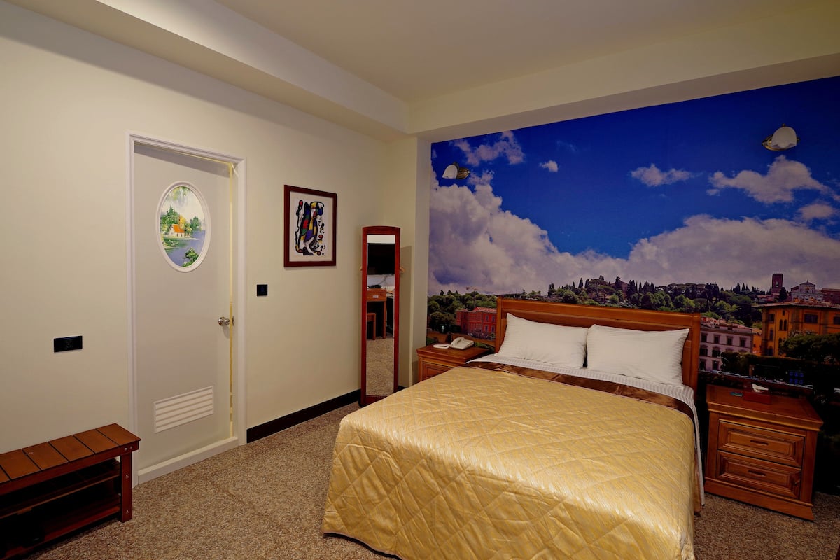 A hotel room with a large bed, a nightstand, a wooden bench, a door with a mirror, and a mural of a cityscape on the wall.