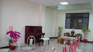 Une salle de conférence avec un piano, des fauteuils à motifs floraux, une table recouverte d’une nappe à carreaux, des plantes en pot et des stanchions rouges.