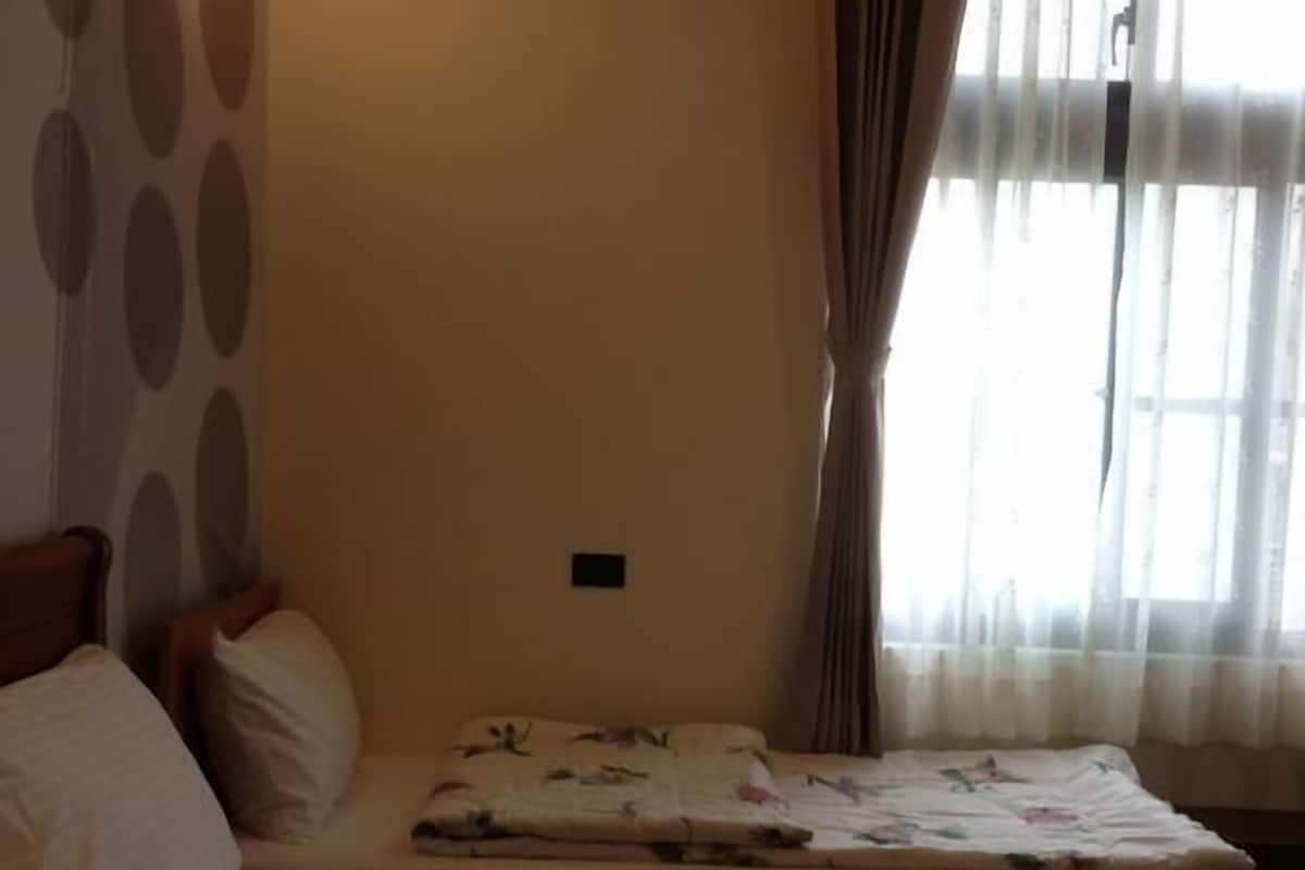 A hotel room with a large bed, a smaller bed, a nightstand, a wall-mounted lamp, and a window with curtains.